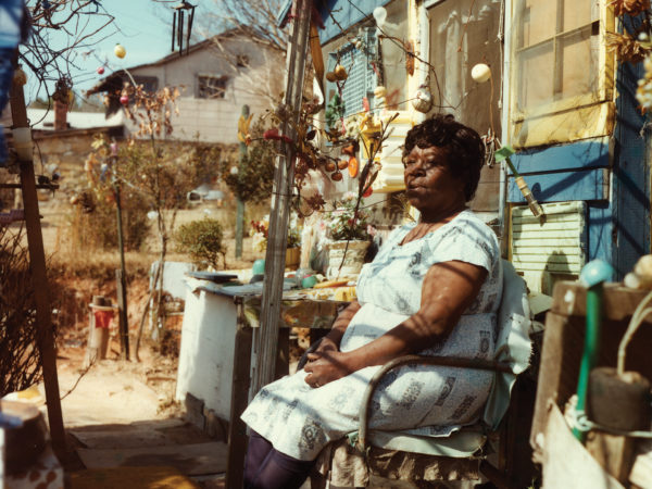 Nellie Mae Rowe sits in a chair on the porch in front of her house under an arbor. The yard in the background contains several small trees covered with multi-colored ornaments.