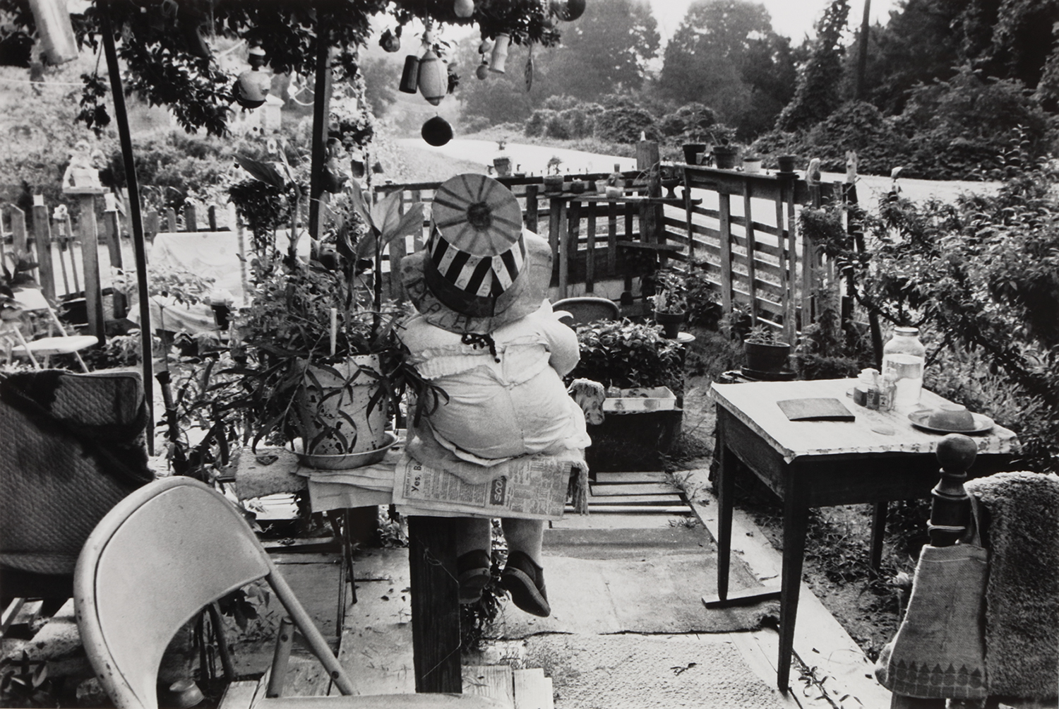 A large clothed doll, wearing a striped top hat and shoes, sits on a bench next to a potted plant with its back facing the camera in the courtyard of Nellie Mae Rowe's garden in this black and white photo. The courtyard is full of mismatched chairs and a table, and a variety of ornaments hang from the tree branches above the doll's head. The doll faces the yard's wooden fence which is topped with potted plants. Beyond the fence, a two-lane road runs in front of the yard, beyond which is a landscape covered with kudzu vines.