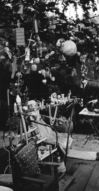 Two chairs with patterned cushions sit in a garden under a small tree. A variety of objects decorate the tree branches including a doll, a teacup, and plastic fruit. Between the two chairs stand some small decorative shelves and a metal rail covered with more objects such as toys, doll heads, and a double strand of pearls.