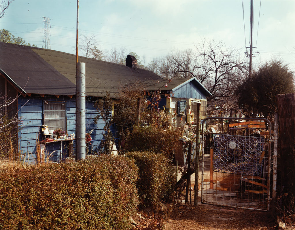 A blue-sided house stands behind a row of bushes and a tall wire fence with a metal gate at the right. A table covered in pots, plants, and decorative objects stands in front of a window. More objects adorn the front of the house and hang from trees and from a wire running next to the house.