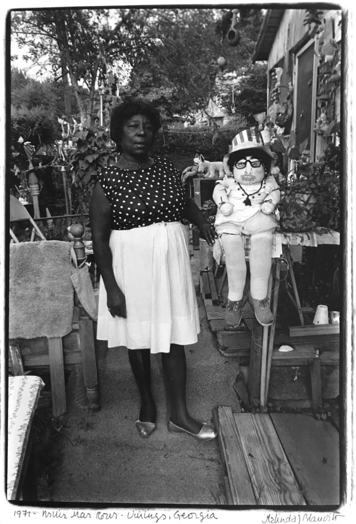 Nellie Mae Rowe stands in the garden next to her house wearing a black shirt with white polka dots and a white skirt. Her hand rests on a table next to a stuffed doll wearing a striped hat, glasses, and shoes.