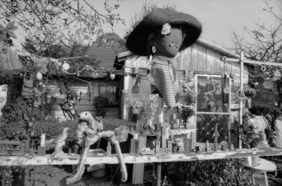 A black and white photograph of the front of Nellie Mae Rowe's house depicts the variety of objects decorating her front yard. A large doll's head wearing a straw hat and a striped dickie stands atop an ornament-covered wire in front of the house. Below it, a wooden rail is covered with a collection of objects including a toy car, a ceramic bird, building blocks, and a dog made of pieces of plastic. Many more objects hang from tree branches and wires in front of the house.