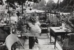 Nellie Mae Rowe’s front yard. A stuffed doll in a white dress and striped top hat leans against a flowerpot planted with diverse foliage. Ornaments hang from a tree above.